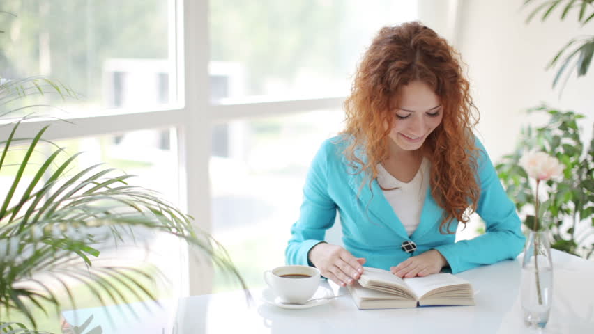 Pretty young woman sitting at table with cup of coffee reading book and smiling