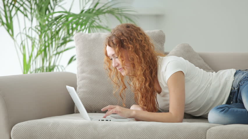 Pretty young woman lying on sofa using laptop and smiling
