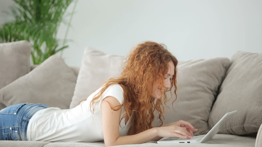 Pretty young woman relaxing on sofa using lapton and smiling at camera
