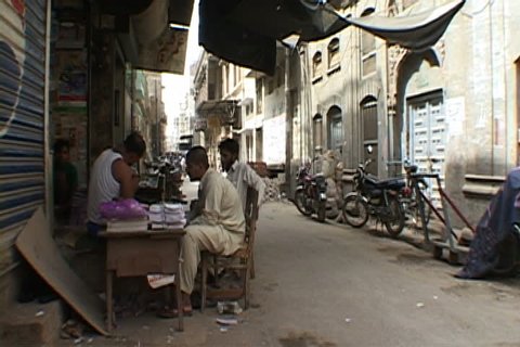 FAISALABAD, PAKISTAN - SEPTEMBER 13, 2002: Men sitting in merchant stall in quiet alley, a motor bike and pedestrians pass by.