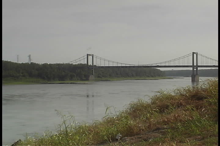 View of the Missouri River under overcast sky. The Paseo suspension bridge crosses the river in the distance in Kansas City, Missouri.