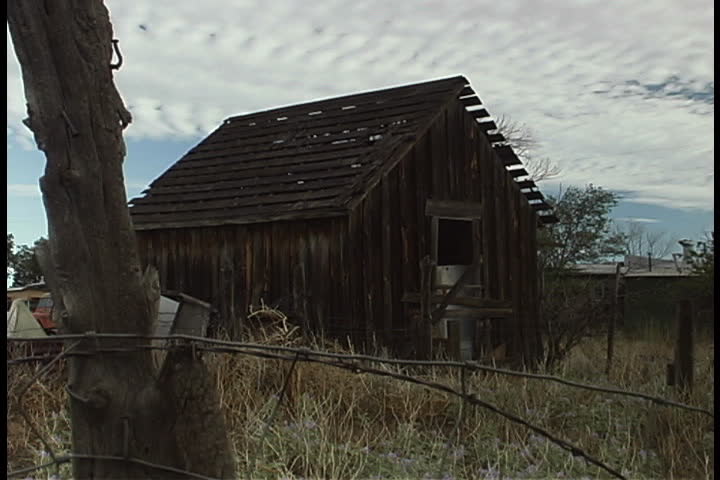 Small, abandoned wooded house with no glass in windows surrounded by tall grass. Barbed wire and remains of fence in foreground in Albuquerque, New Mexico.