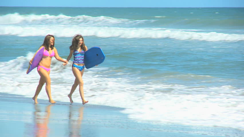 Pretty young teenage girls running along beach by ocean carrying body board