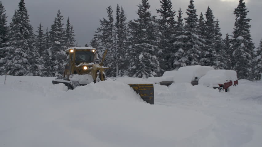 Large road grader with plow clears snow off buried vehicles after heavy