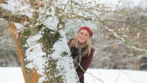 Beautiful young blonde female behind tree playfully throwing snow on winters day