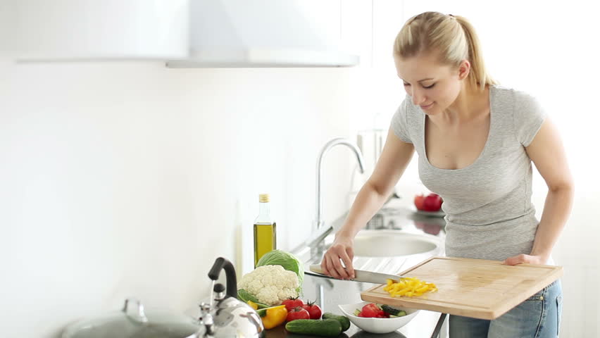 Young woman in kitchen adding fresh cut bell peppers into salad

