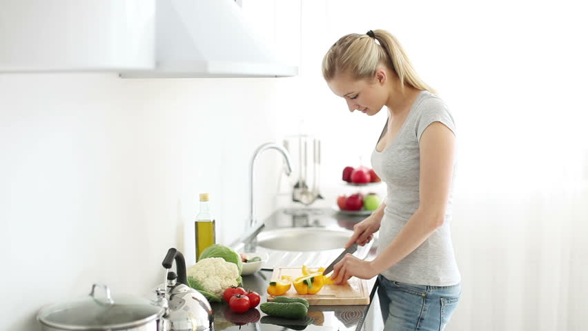 Smiling young woman in kitchen cutting bell peppers on cutting board for salad
