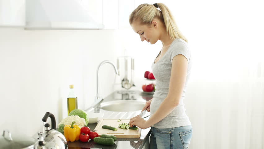 Smiling young woman standing at kitchen table cutting cucumbers on cutting board