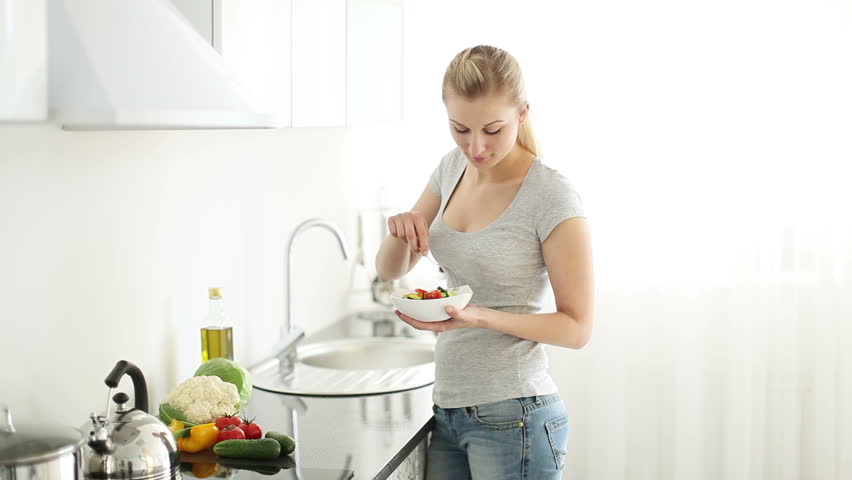 Pretty young woman standing in kitchen eating vegetable salad and smiling at
