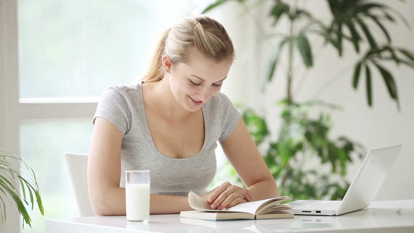 Young woman sitting at table reading book drinking milk and smiling at camera
