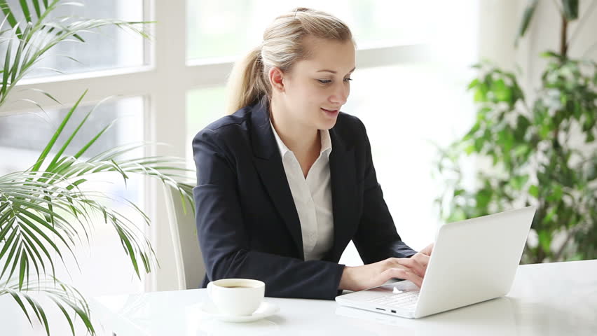 Smiling young woman at office sitting at table using laptop and smiling at