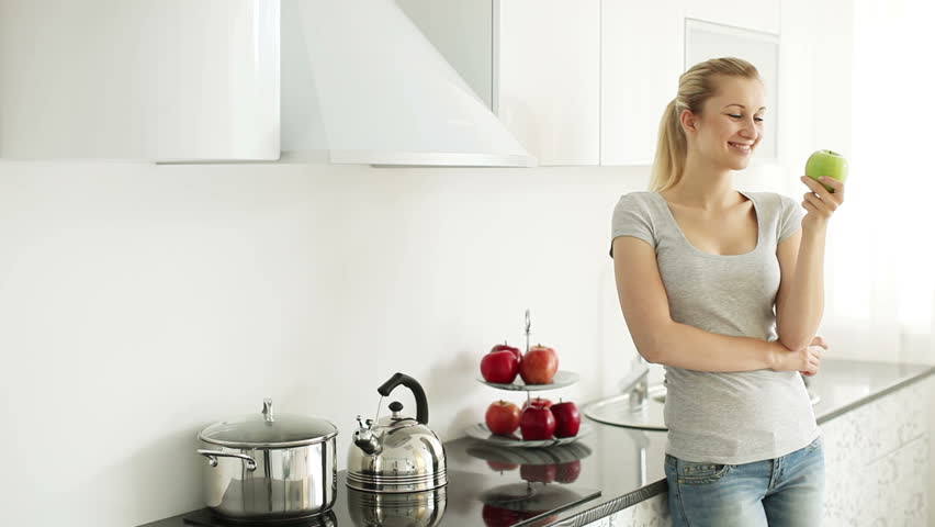 Young woman standing in kitchen eating green apple and smiling
