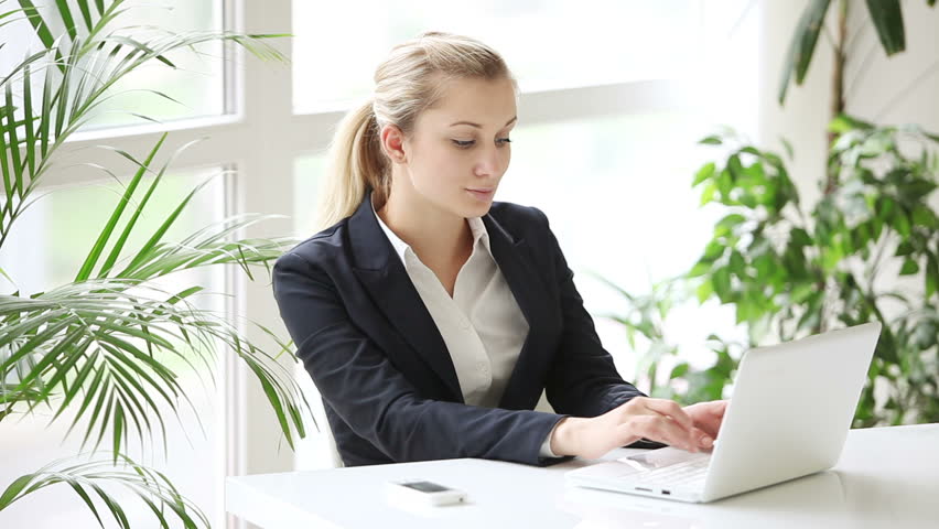 Young woman dressed in formal wear sitting at table at office and using laptop
