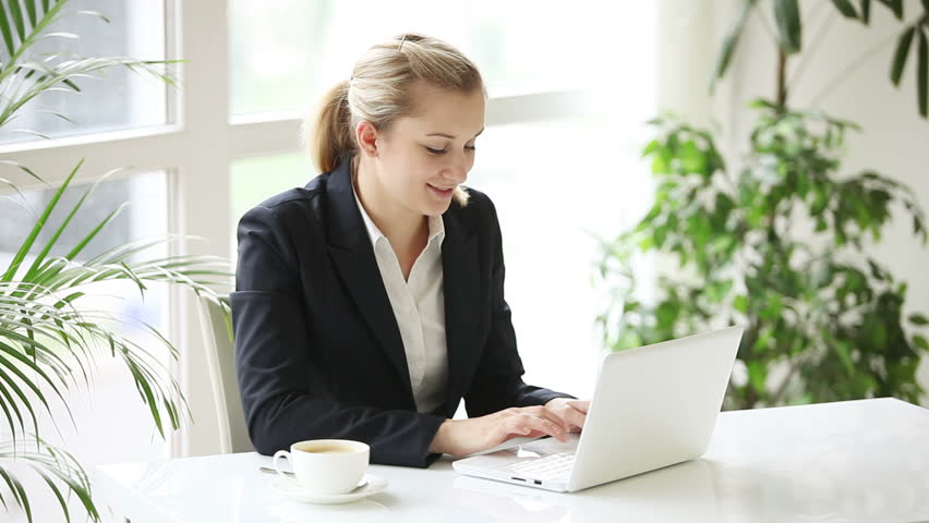 Young business woman sitting at office table with cup of coffee using laptop and