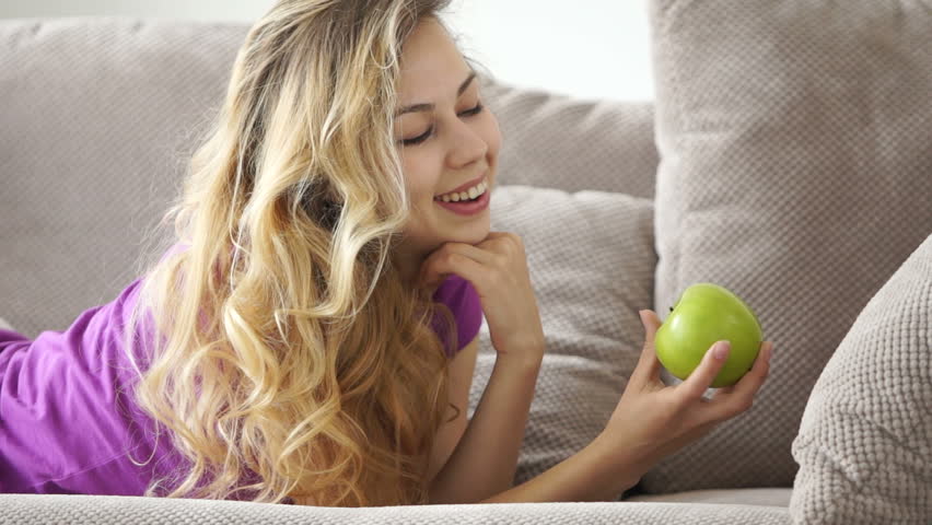 Pretty young woman on sofa holding apple smiling at laughing at camera
