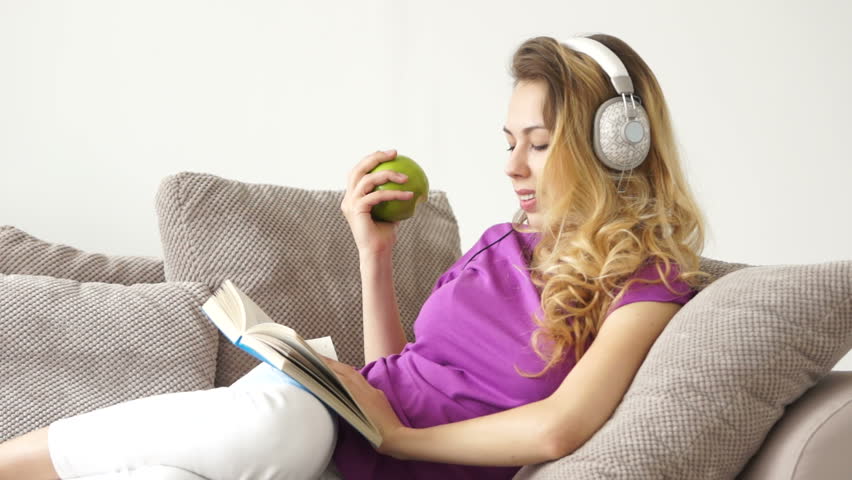 Pretty young woman lying on sofa listening to music on headphones reading book
