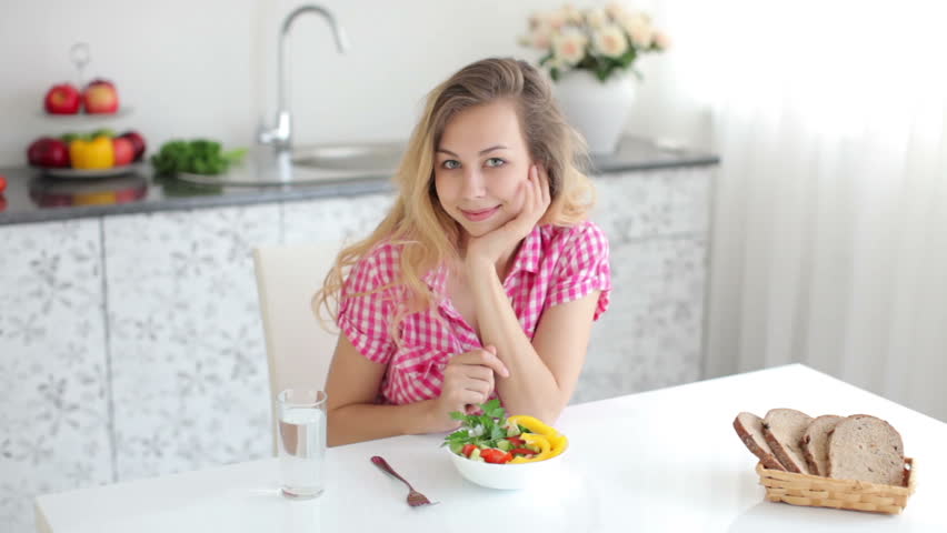 Pretty young woman sitting at table in kitchen and eating vegetable salad with