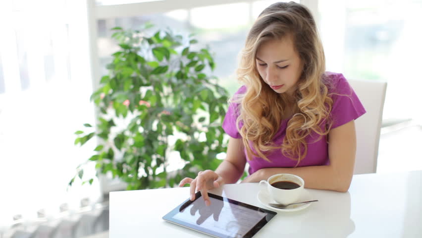 Pretty young woman sitting at table with cup of coffee using touchpad smiling