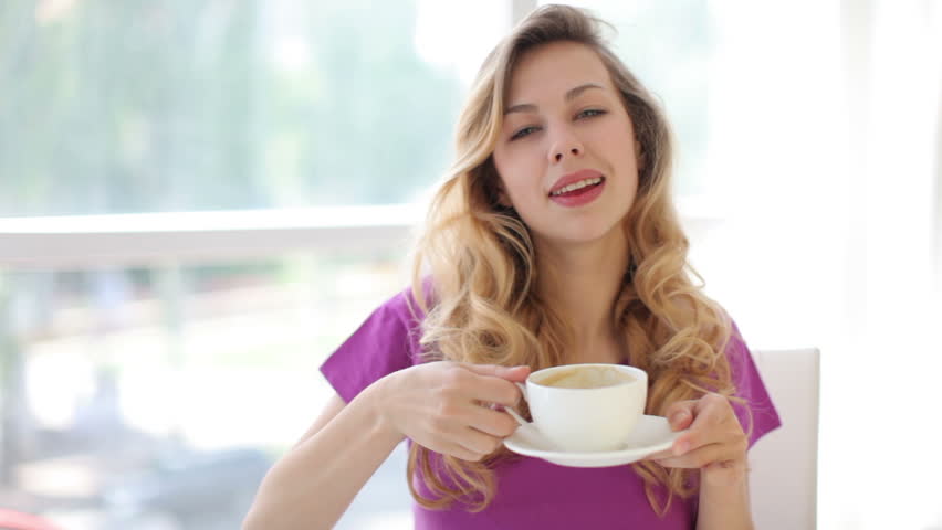 Pretty young woman sitting at table enjoying coffee and smiling at camera

