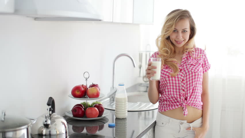 Pretty girl standing in kitchen with glass of milk and laughing
