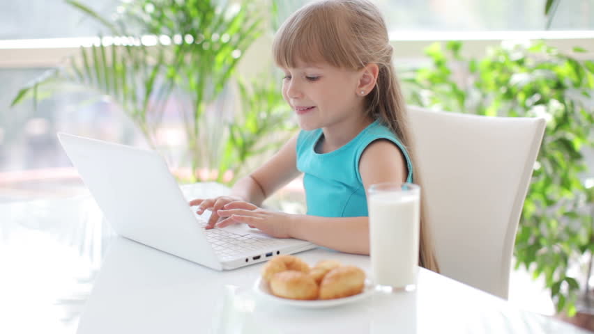 Cute little girl sitting at table with glass of milk and plate of cakes using