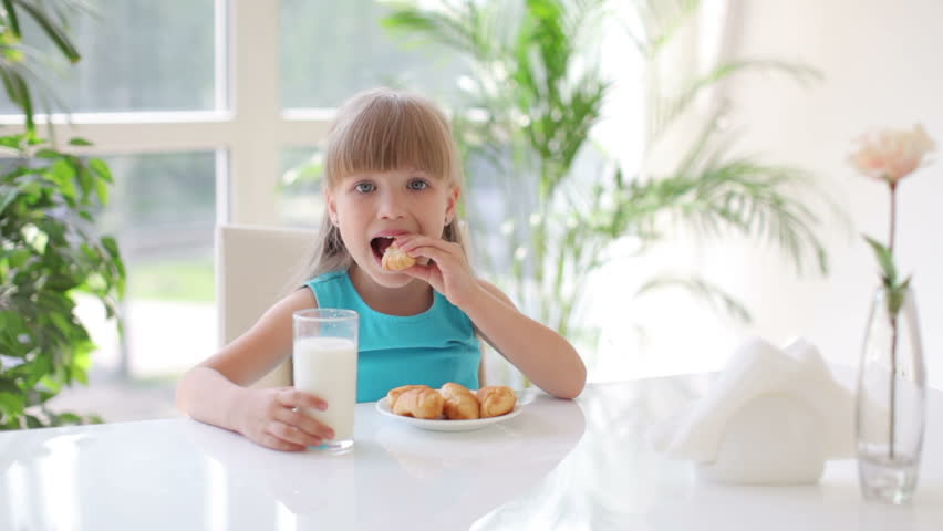Cute little girl sitting at table eating cakes and drinking milk and laughing at