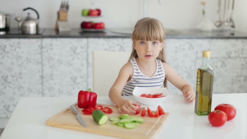 Cute little girl sitting at kitchen table and putting fresh-cut vegetables into