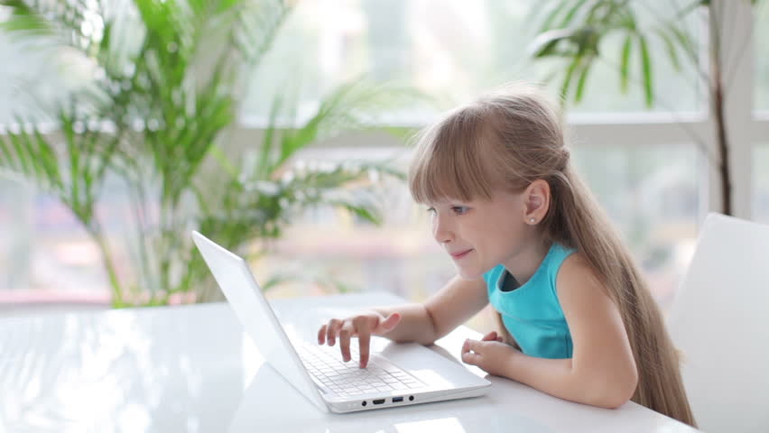 Beautiful little girl sitting at table using laptop and smiling at camera
