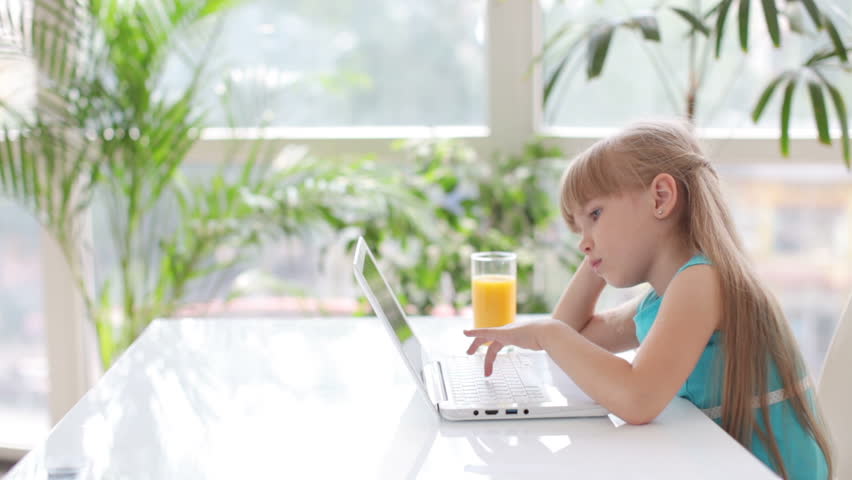 Beautiful little girl sitting at table using laptop and looking thoughtfully out