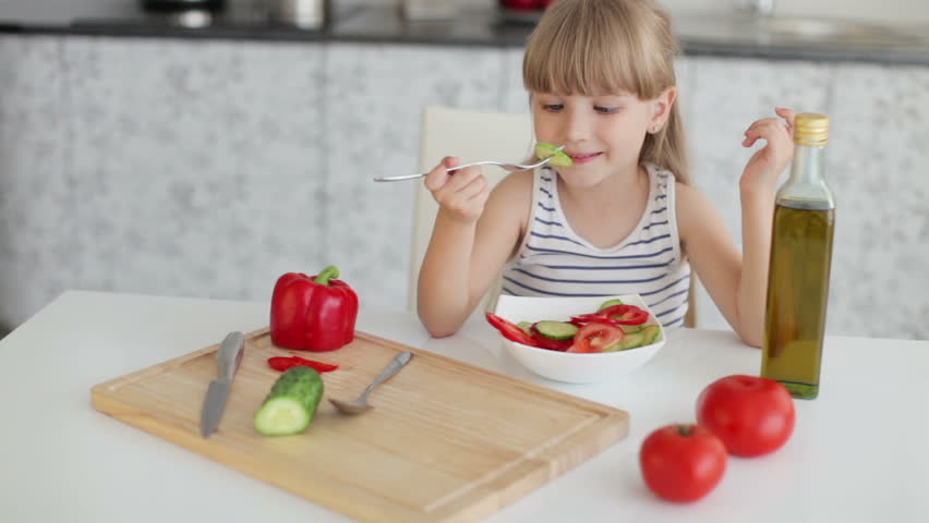 Cute little girl sitting at kitchen table eating vegetable salad and smiling at