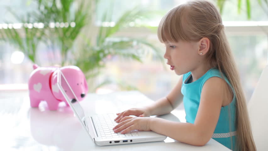 Cute little girl sitting at table with laptop smiling and laughing at camera
