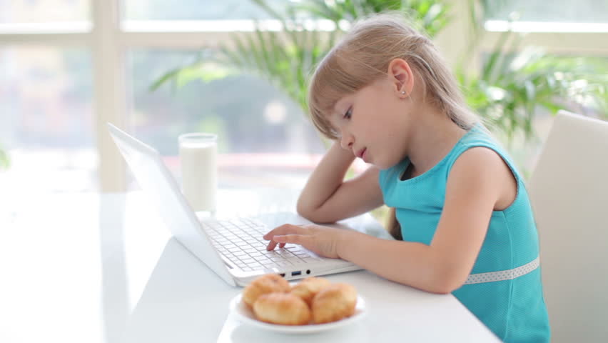Beautiful little girl sitting at table leaning on her hand and using laptop
