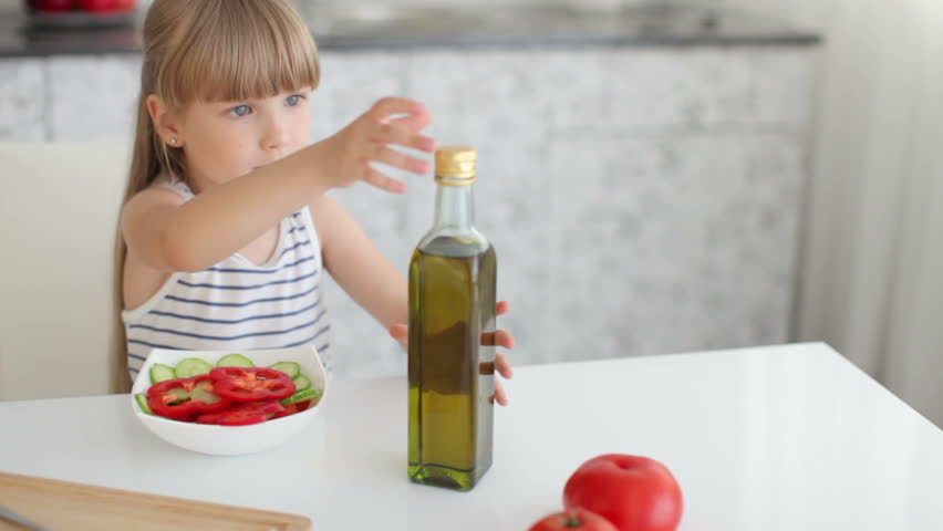 Cute little girl sitting at kitchen table and pouring olive oil from bottle into