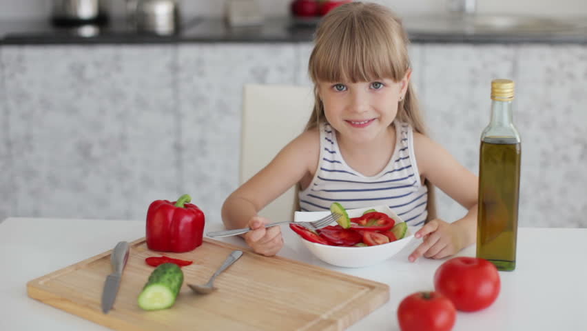 Funny little girl sitting at kitchen table and eating vegetable salad from bowl
