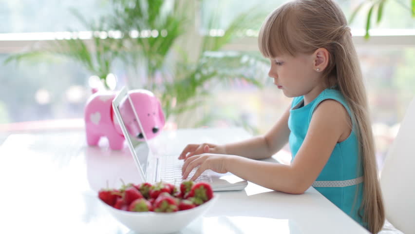 Cute little girl sitting at table with bowl of strawberries and using laptop
