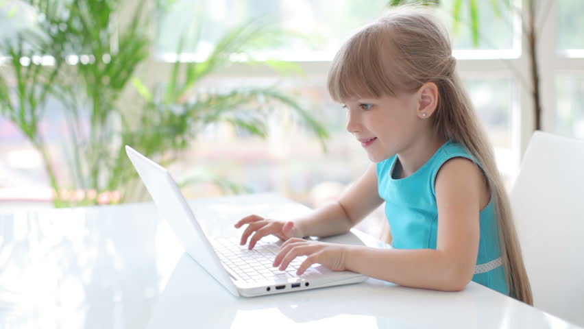 Cute little girl sitting at table with laptop smiling and showing thumb up at