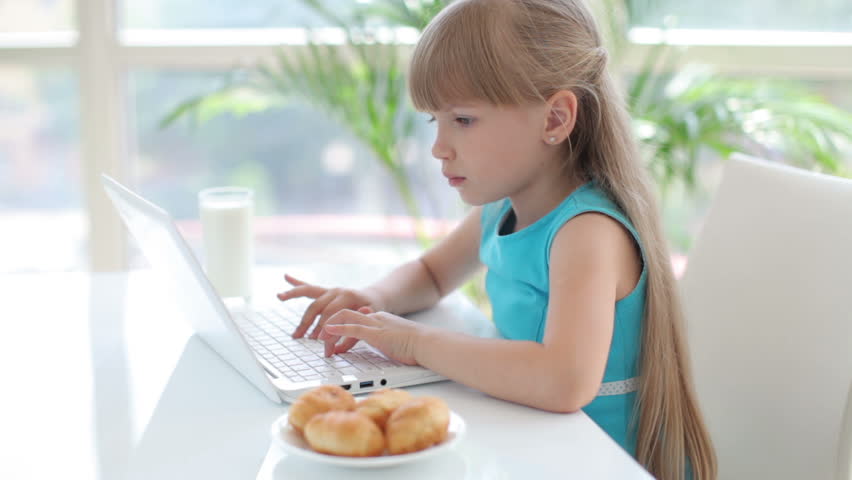 Cute smiling little girl sitting at table using laptop and looking at camera
