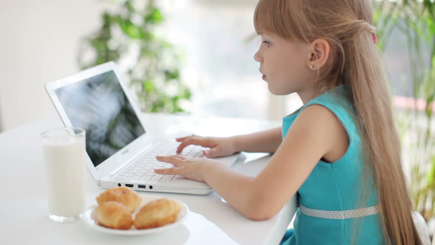 Cute little girl sitting at table eating cakes and using laptop
