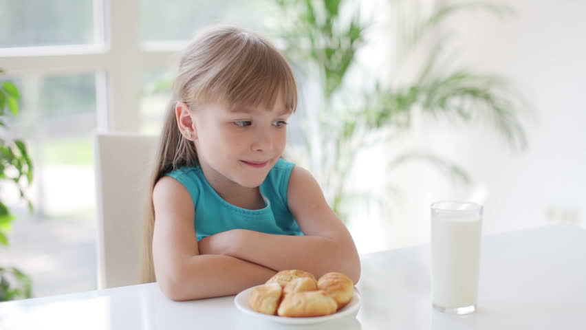 Cute little girl sitting at table with plate of cakes and glass of milk in front
