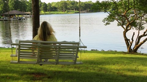A blonde haired woman enjoys swinging on a wooden swing that over looks a sparkling lake on a summer day