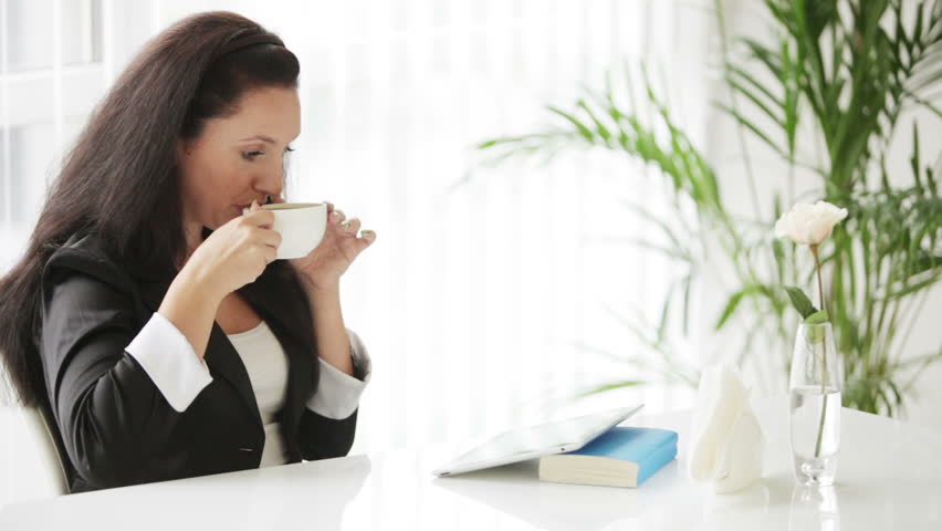 Pretty young woman sitting at table using touchpad holding cup of coffee and
