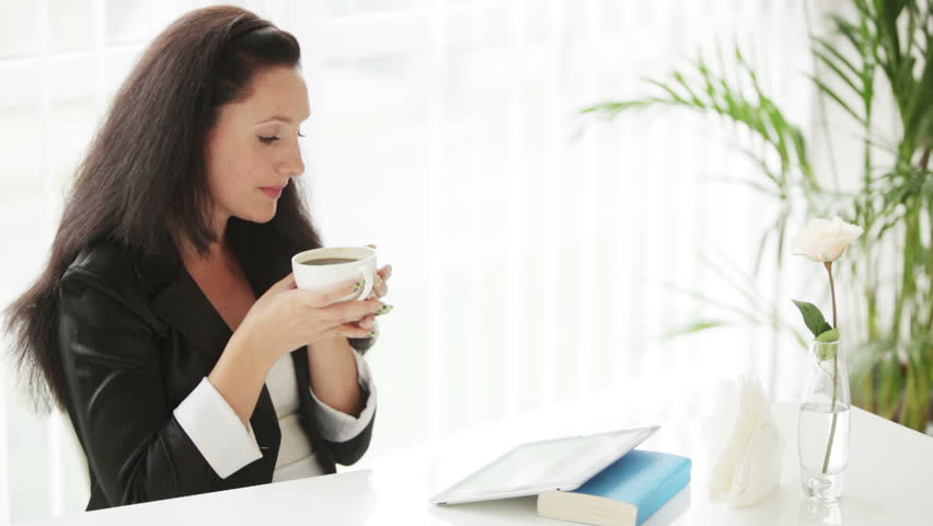 Young woman sitting at table drinking coffee using touchpad smiling and looking
