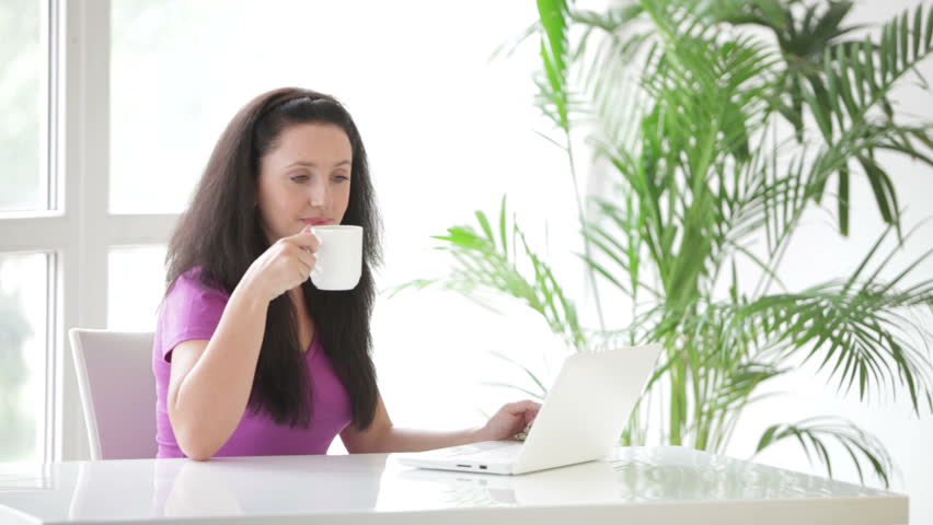 Pretty young woman sitting at table using laptop drinking coffee and smiling at