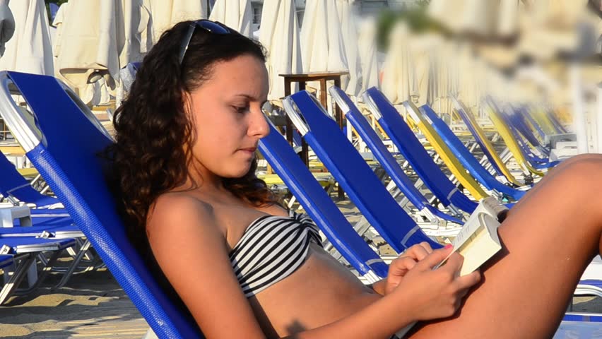Student teenager girl reading a book on a beach while sunbathing.