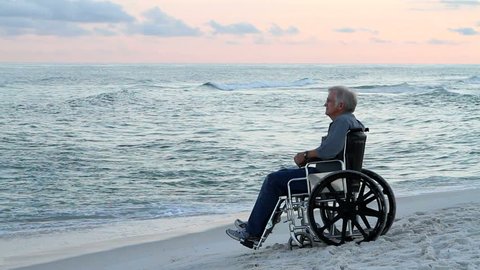 Elderly senior, physically disabled man sits in a wheelchair at the seashore watching the waves roll in and thinking.