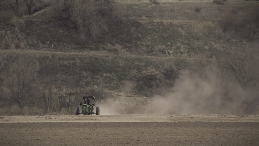 Farm worker drives an old tractor over a dry agricultural field, kicking up a