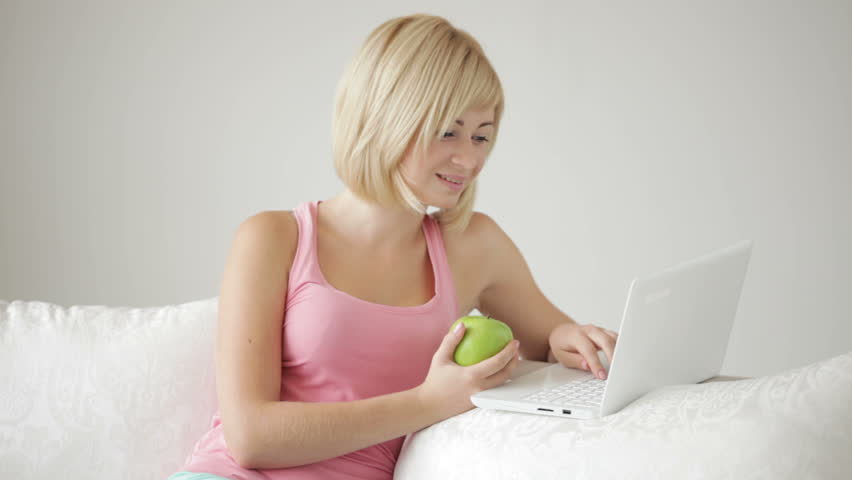 Attractive young woman sitting on sofa using laptop biting apple and smiling
