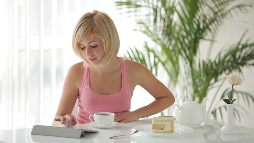 Attractive young woman sitting at table with touchpad drinking tea eating cake