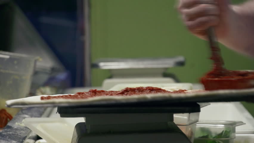 Male chef liberally spreads tomato sauce over gourmet pizza pie with a large