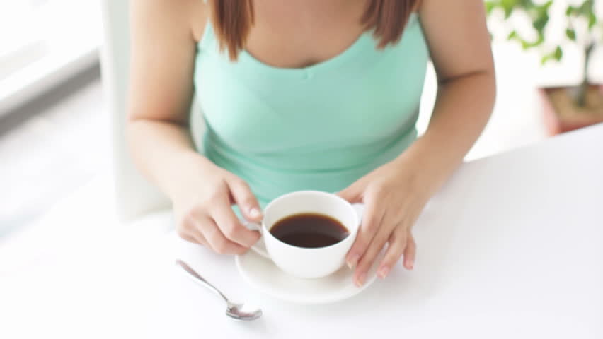 Pretty girl sitting at table holding cup of coffee and smiling at camera

